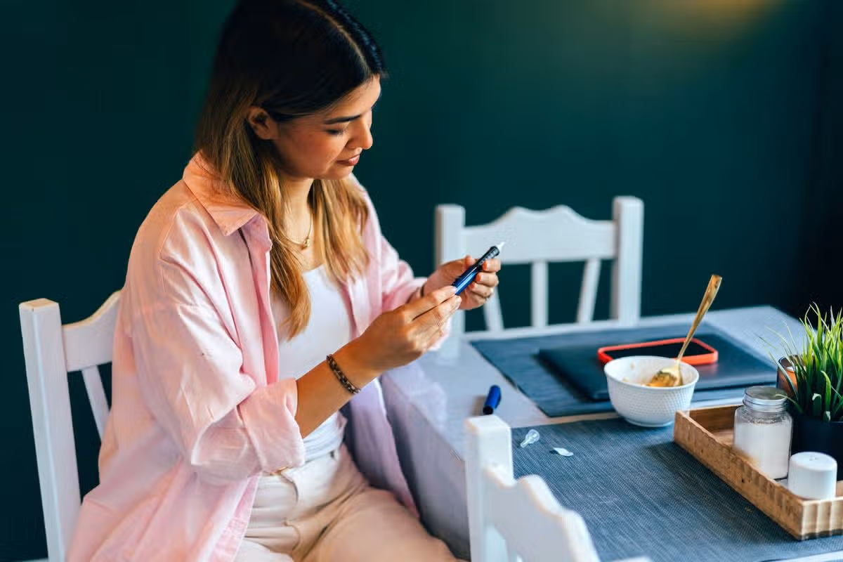 An individual is seated at a table, engrossed in their mobile device while wearing a pink long-sleeved top. The table is set with a bowl, a plant in a small pot, and a tray with a cup and a saucer. Behind them is a wall painted in a dark green hue, and adjacent to the table, there is a white chair.