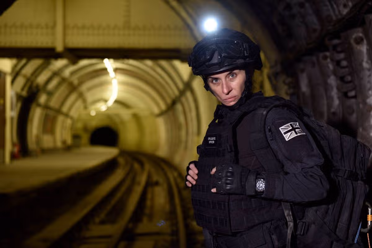 An individual dressed in a black tactical uniform, complete with a helmet and a backpack, is positioned in a dimly lit tunnel with visible railway tracks extending into the distance.