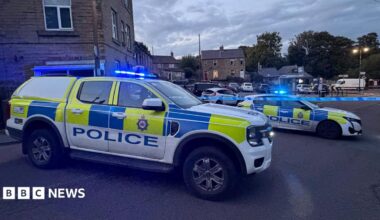 Two police cars with lights flashing parked alongside police cordon tape.