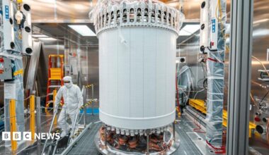 The LUX-ZEPLIN main detector in a laboratory. The LZ is a large white cylindrical machine. A person wearing a white hazmat suit is scene walking down some stairs next to the machine.