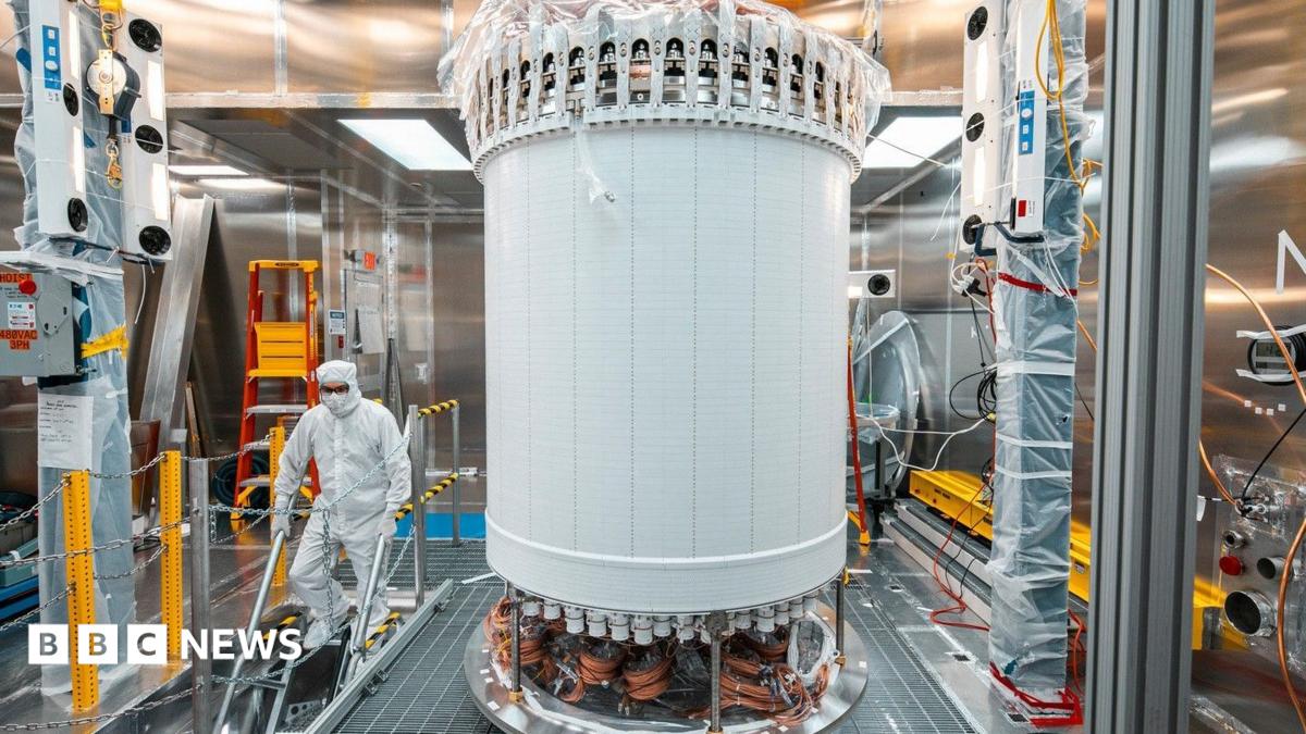 The LUX-ZEPLIN main detector in a laboratory. The LZ is a large white cylindrical machine. A person wearing a white hazmat suit is scene walking down some stairs next to the machine.