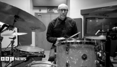 A black-and-white image of a bald man wearing glasses and a dark shirt sitting behind a drum set with drum sticks in hand.