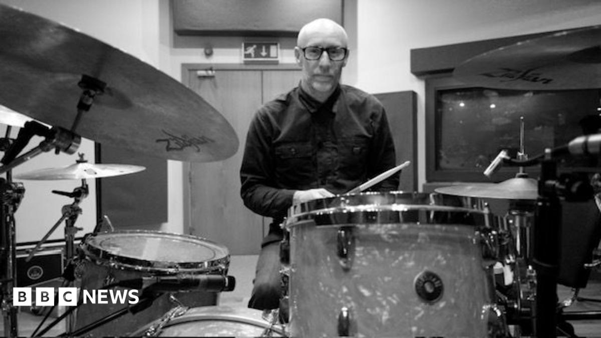 A black-and-white image of a bald man wearing glasses and a dark shirt sitting behind a drum set with drum sticks in hand.