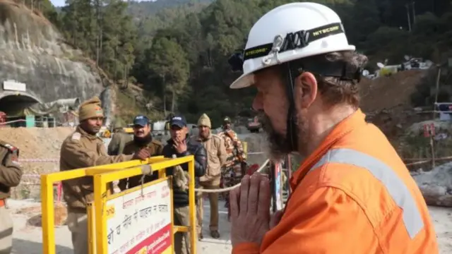 Arnold Dix is pictured wearing an orange hi-viz jacket and white safety helmet, in a mountain area.
