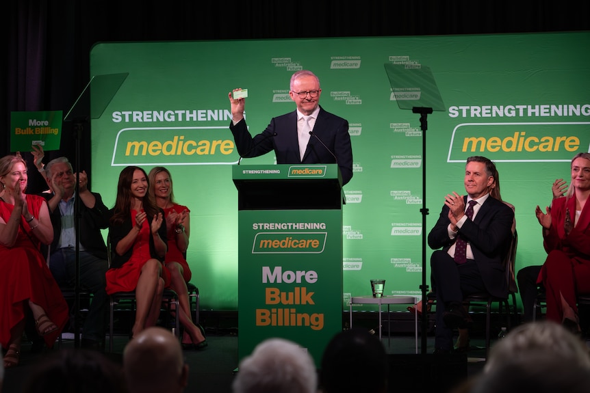 Anthony Albanese on stage, holding up a Medicare card, as people clap.