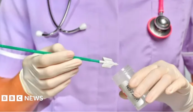 A woman in white rubber gloves holds a test in one hand and a pot to deposit it in the other.  A stethoscope hangs round her neck and she wears a purple uniform.