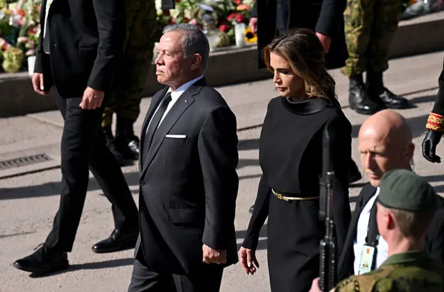 King Abdullah II, who has grey hair, wears a black suit, whit shirt and black tie, while Queen Rania, who has brown long hair, wears a black dress