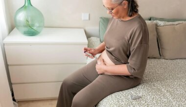 A photo of a mature woman sitting on the edge of a bed and self injecting in her abdomen.