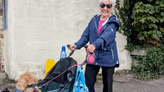 Barbara Ramsay is standing holding a pram which has a small brown dog in it. She is smiling and wearing sunglasses.