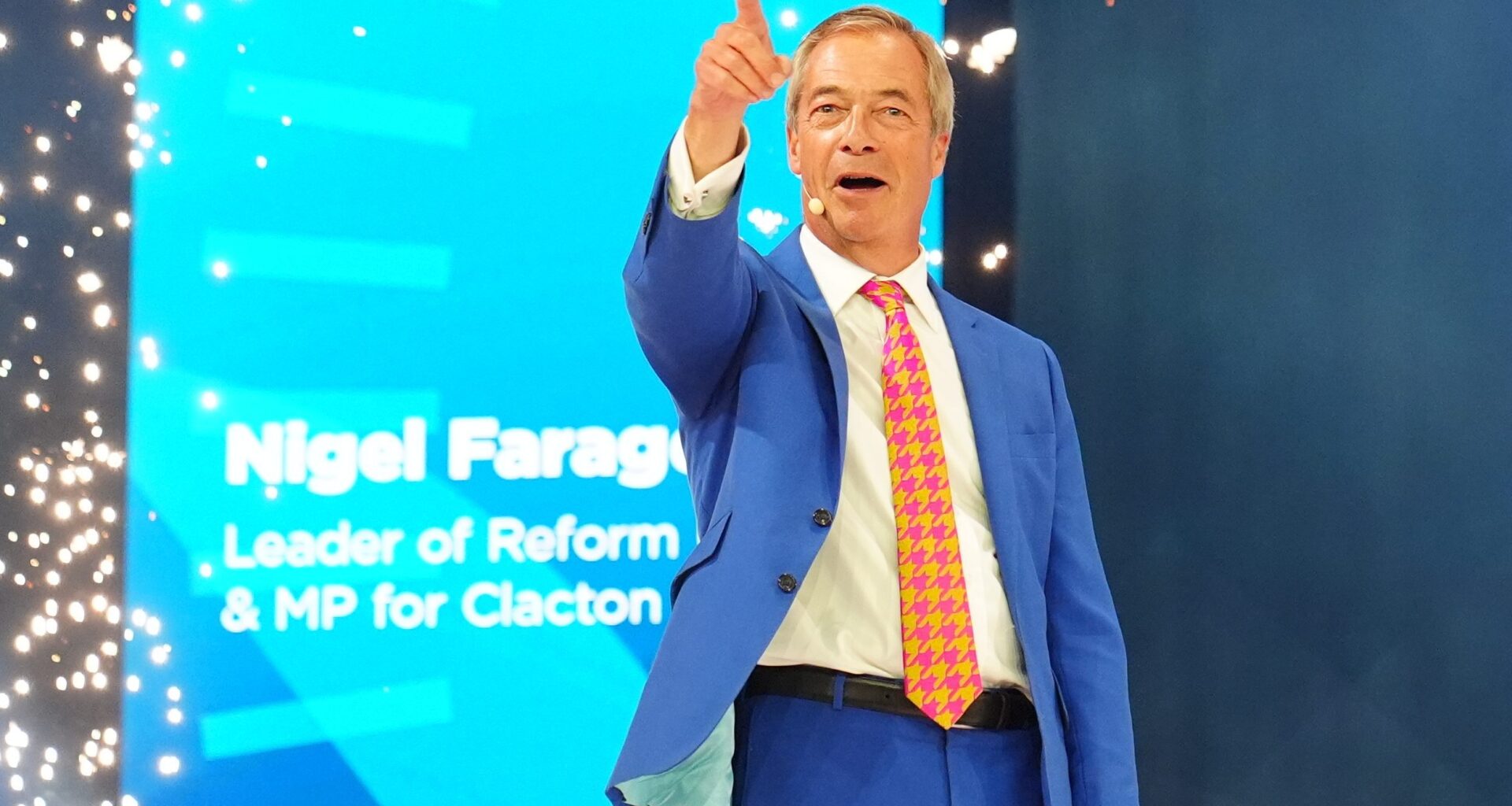 Nigel Farage speaking at the Reform UK conference, pointing upwards with a confident expression.