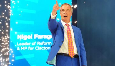 Nigel Farage speaking at the Reform UK conference, pointing upwards with a confident expression.