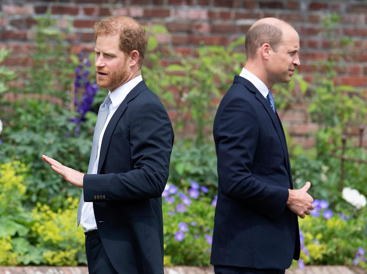 Two people stand with their backs to one another in a garden in front of a red brick wall.