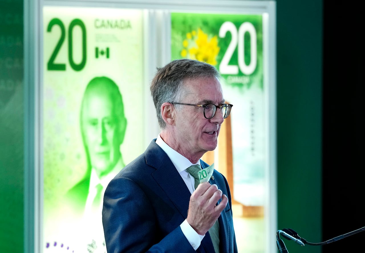 A person stands at a microphone in front of a picture of Canada's new $20 note with a portrait of King Charles on it.
