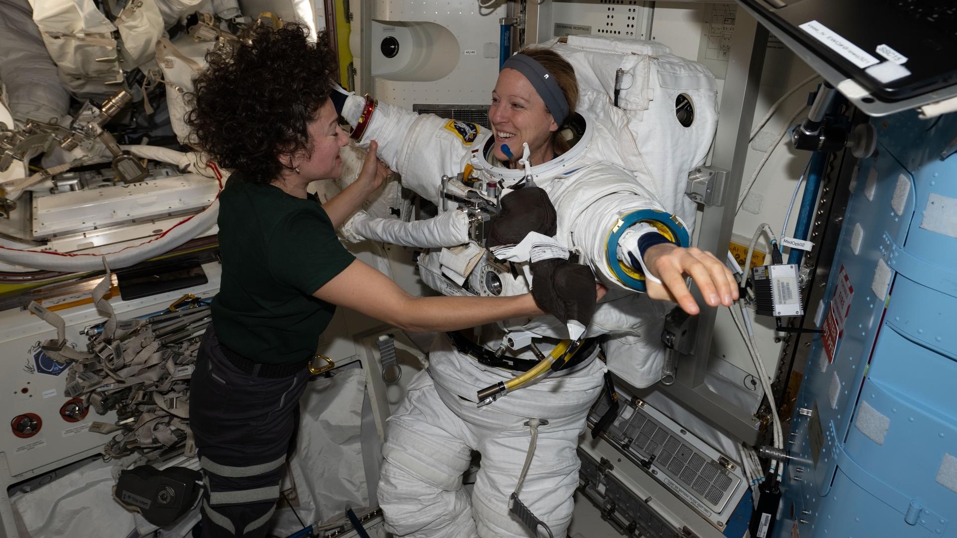 a woman in a black polo shirt helps a smiling woman put on a white spacesuit aboard the space station