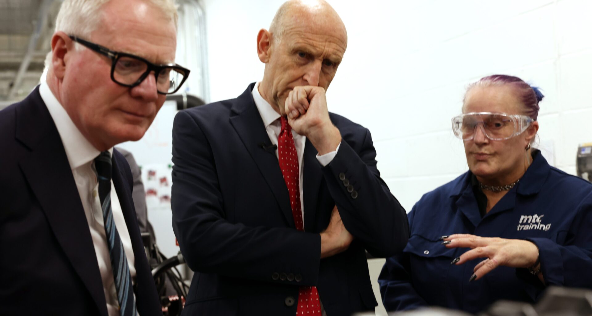 Two men in suits and a woman in safety glasses and workwear at a factory.