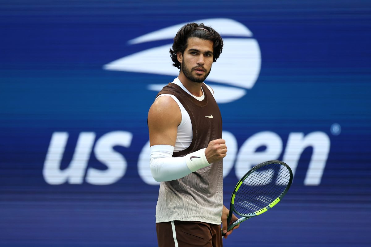 Tennis player in brown and white athletic wear, holding a racket, fist clenched, US Open background