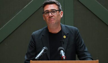 Andy Burnham speaking at a press conference, addressing current events and regional developments, wearing a suit and tie.