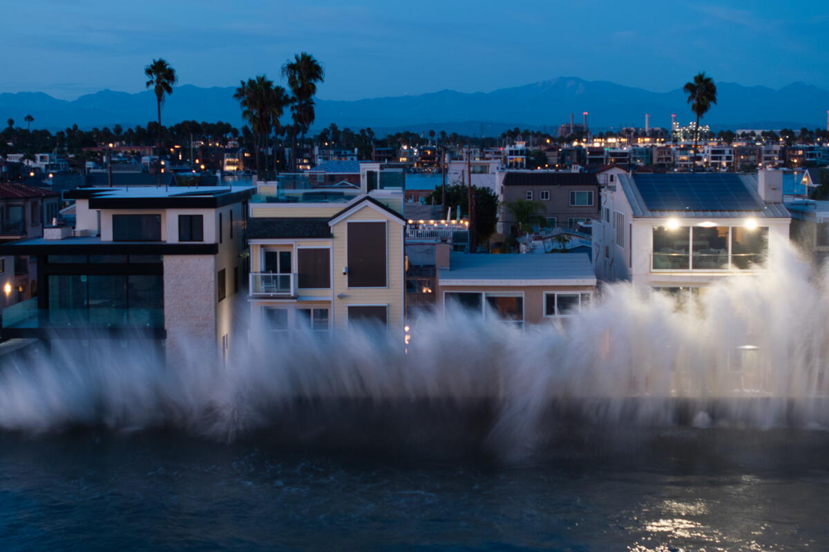 A large ocean wave hits a row of close-set, multistory homes at nightfall.