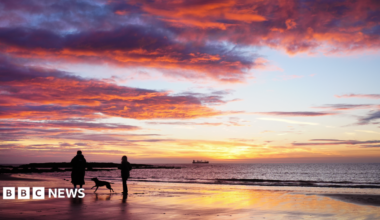 Dog walkers at sunrise on Tynemouth beach in the North East of England. There are two people and a dog running between them. Beyond them the sky is yellow and blue and the clouds bright orange.