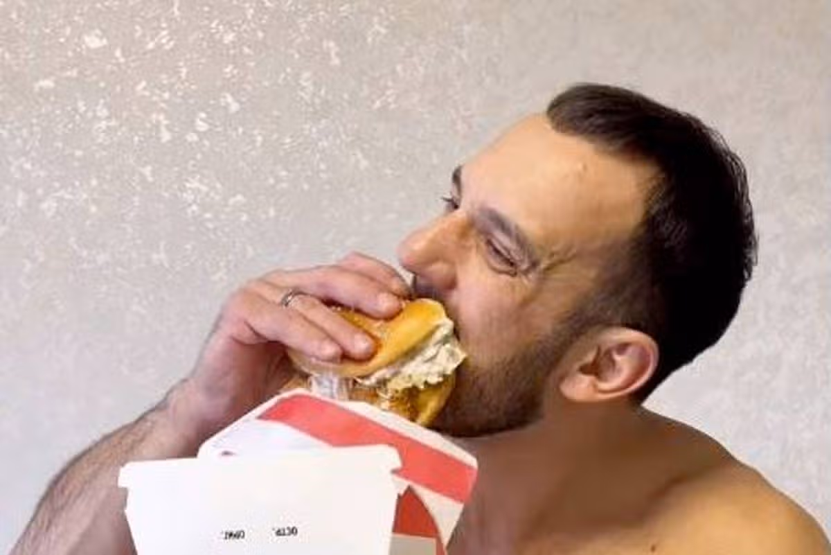 An individual is engaged in the act of consuming a sandwich from a box, displaying a focused expression. The sandwich features a combination of various ingredients.