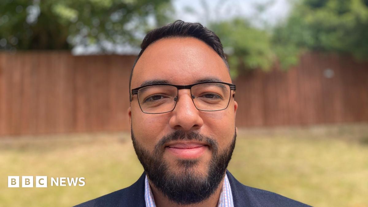 Man wearing a blue suit and checked shirt looks at the camera. He is wearing black glasses and has black hair and a beard.