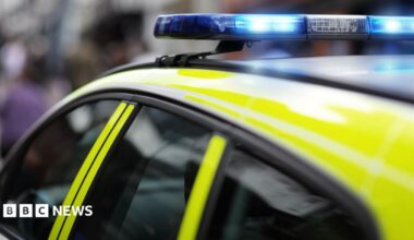 The roof of a police car with bright yellow markings and a lit blue emergency light bar, parked on a street with blurred buildings and people in the background.