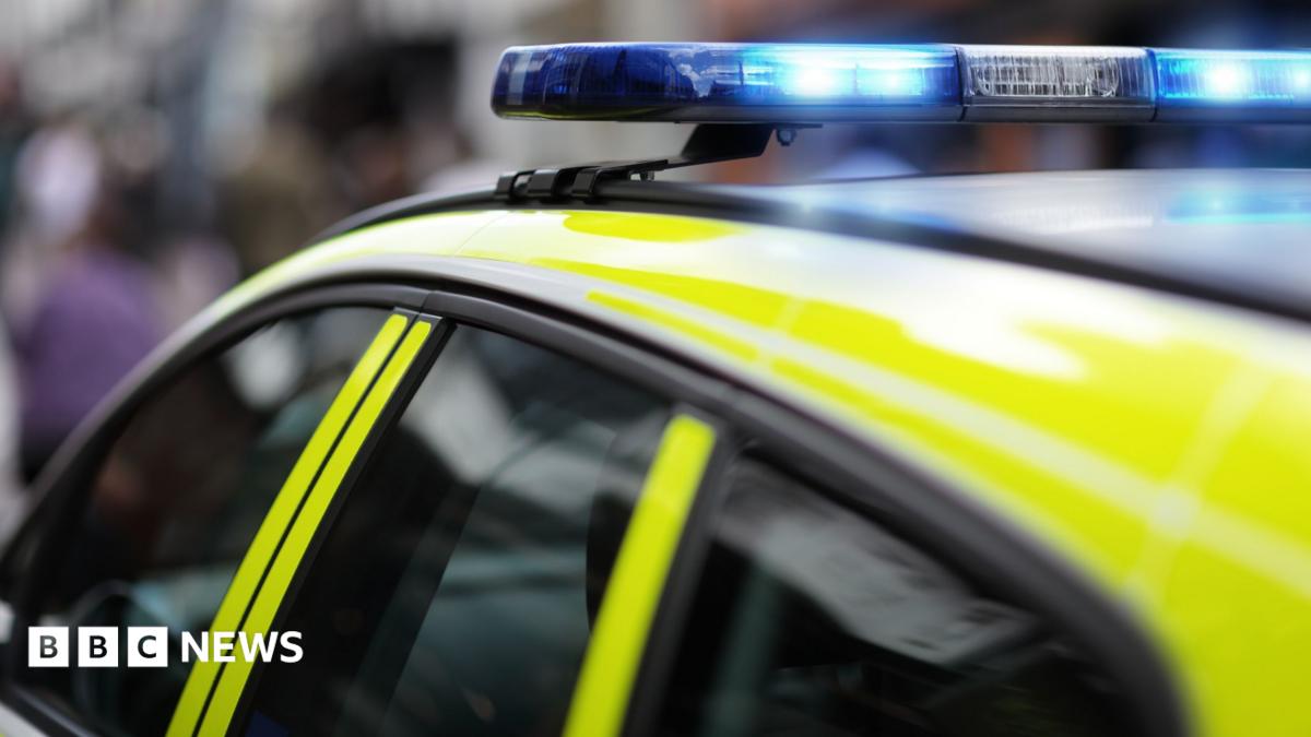 The roof of a police car with bright yellow markings and a lit blue emergency light bar, parked on a street with blurred buildings and people in the background.