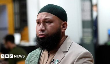 Mothin Ali looking to his right, wearing a beige suit jacket with a green and white scarf draped over his right shoulder