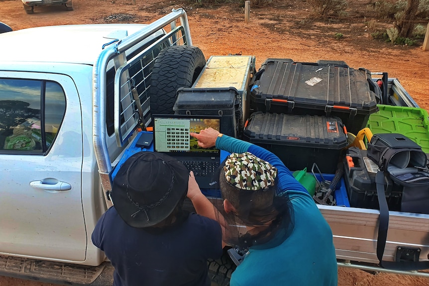A laptop sitting on the back of a ute, with two people looking at the screen