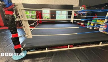 A boxing ring that looks like it has seen better days, with black bin bags wrapped around the poles. It is in a gym with black padded floors, and there are some windows behind it which look out into the forest.