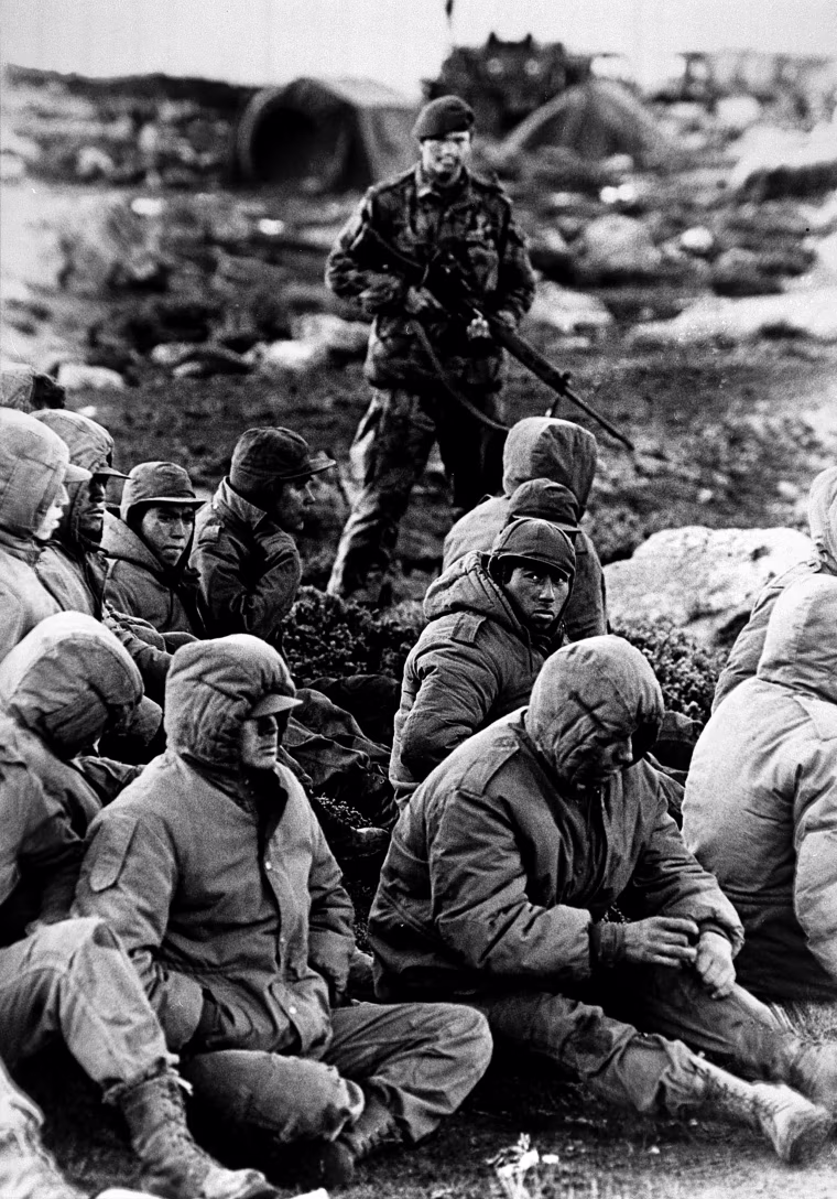 Captured Argentinian soldiers guarded by a British Royal Marine as they await transit out of the area at Goose Green, Falkland Islands, in 1982.