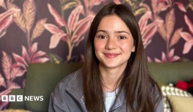 Gala Fraser Lim has long brown hair and is smiling while sitting on a sofa. The wallpaper behind her is brown with pink flowers on it.