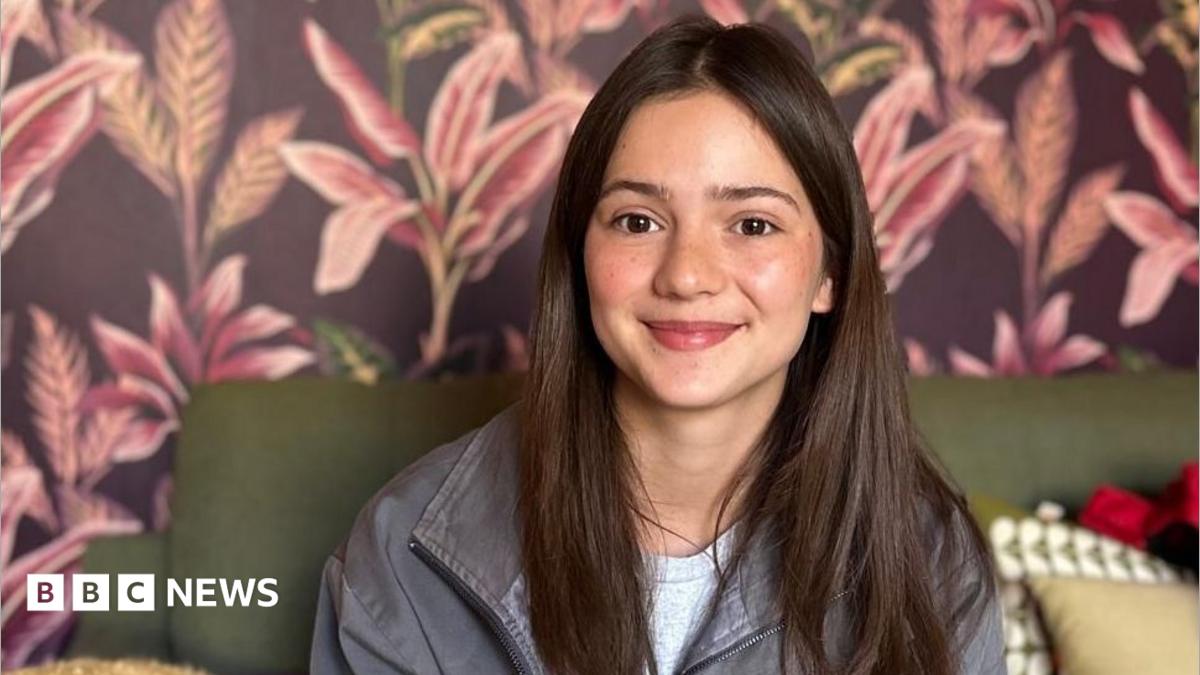Gala Fraser Lim has long brown hair and is smiling while sitting on a sofa. The wallpaper behind her is brown with pink flowers on it.