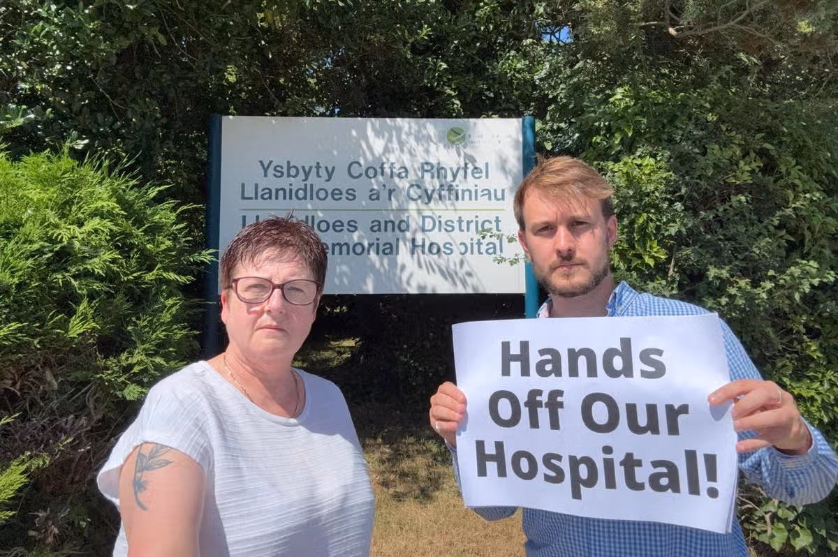 Two people, one with a poster saying 'Hands off our hospital', near a hospital