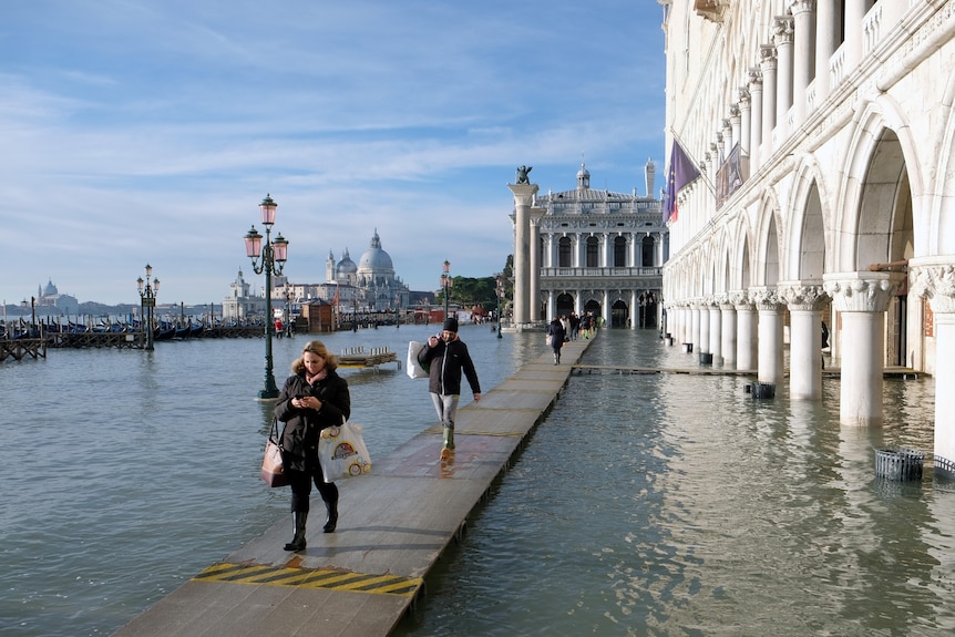 People walk on wooden boards placed across water near buildings.