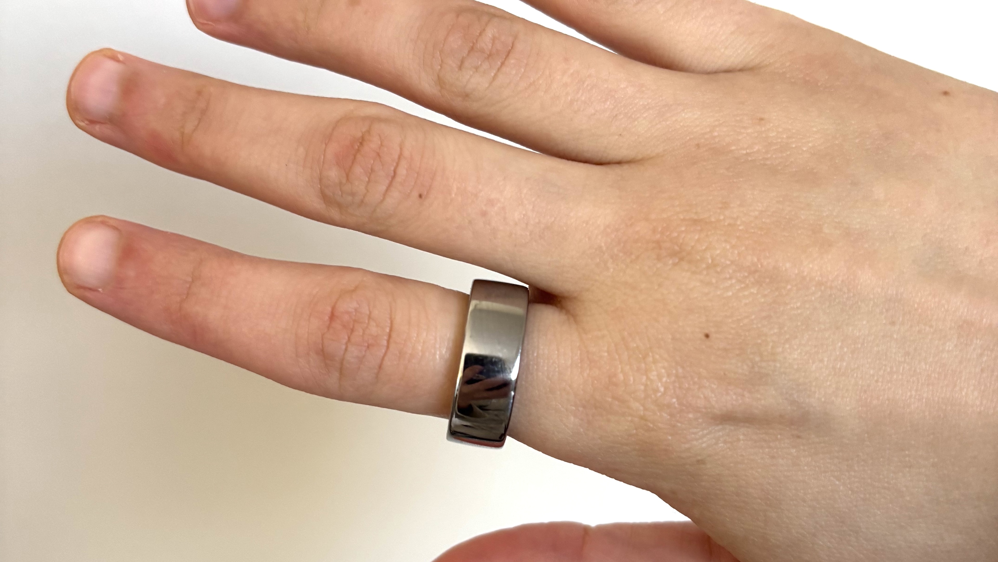 A close-up of the silver Oura Ring 4 on a pointer finger in front of a white wall.
