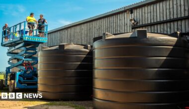 Contractors inspecting two large rainwater harvesting systems on a farm. The systems are two large plastic black containers which are placed to the side of an industrial barn. There is a blue cherry picker to the rear with three workpeople standing on it.