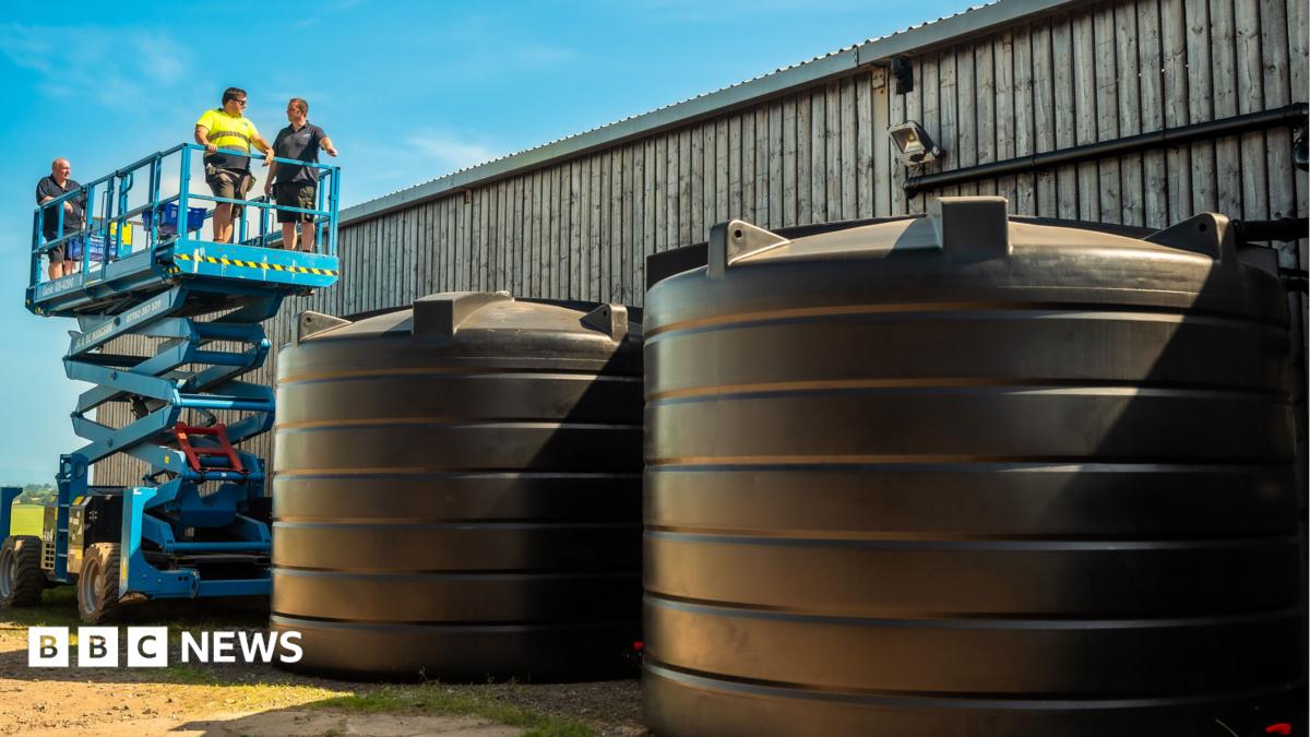 Contractors inspecting two large rainwater harvesting systems on a farm. The systems are two large plastic black containers which are placed to the side of an industrial barn. There is a blue cherry picker to the rear with three workpeople standing on it.