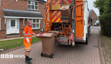 A refuse collector is wearing a hi-vis top, shorts and boots. He is loading two food waste wheelie bins on to a lorry to be emptied.