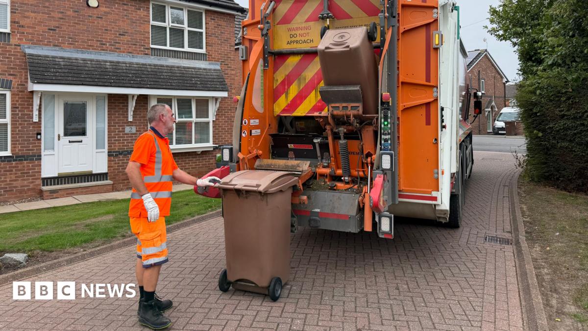 A refuse collector is wearing a hi-vis top, shorts and boots. He is loading two food waste wheelie bins on to a lorry to be emptied.