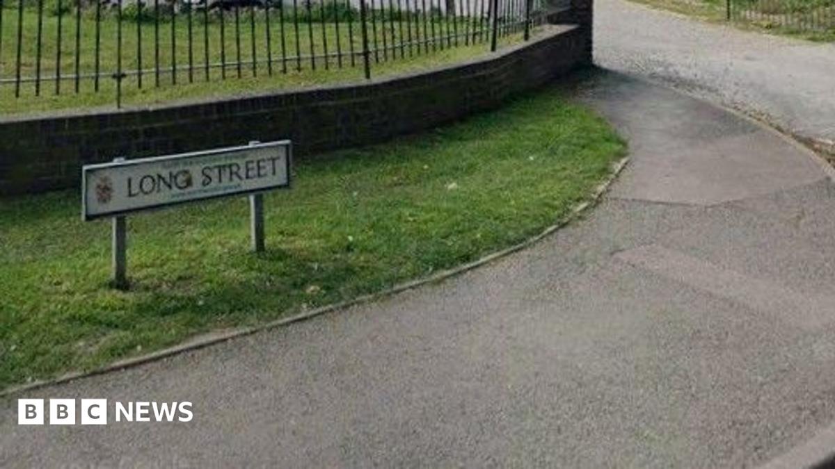 A low street sign with Long street written on it. The G and S look to have been vandalised. the sign stands on grass, there is a pavement in front of it.
