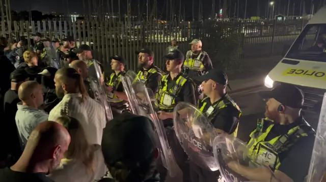 A row of police with riot shields standing in front of protesters at a port in Portsmouth following the rescue of 140 migrants from the Channel