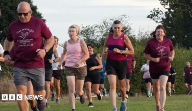 A group of men and women running in a grass field. Some wearing matching maroon coloured T-shirts which are the club colours.