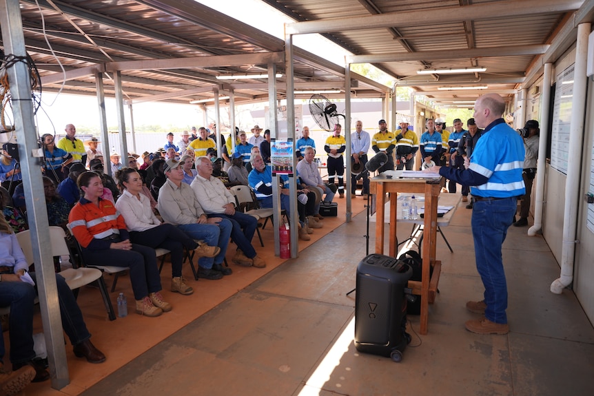 A mine worker in high-vis stands before a seated crowd, speaking about a fracking project.