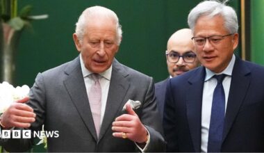 King Charles wearing a grey suit jacket and white shirt and pink tie, talking to Jensen Huang, the CEO of Nvidia, who is wearing a dark suit, white shirt and blue tie. Former Google DeepMind boss Sir Demis Hassabis is walking behind them.