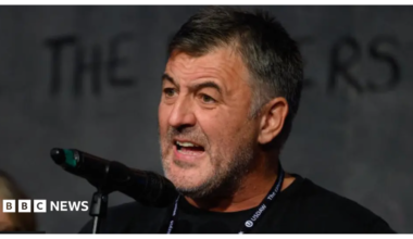 A man with short, black hair and stubble stands in front of a microphone mid-speech. He is wearing a black t-shirt and has a lanyard around his neck
