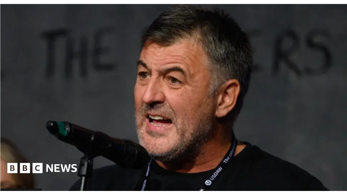 A man with short, black hair and stubble stands in front of a microphone mid-speech. He is wearing a black t-shirt and has a lanyard around his neck