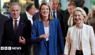 A smiling Mark Carney, wearing a suit, walks alongside European Parliament President Roberta Metsola and Ursula von der Leyen at the European Parliament