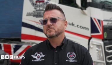 A man wearing sunglasses and a black shirt. He is standing in front of a transportation vehicle.