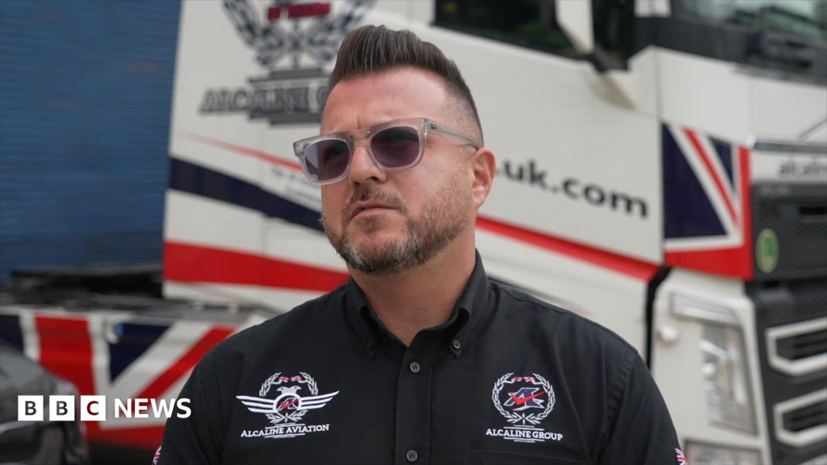 A man wearing sunglasses and a black shirt. He is standing in front of a transportation vehicle.
